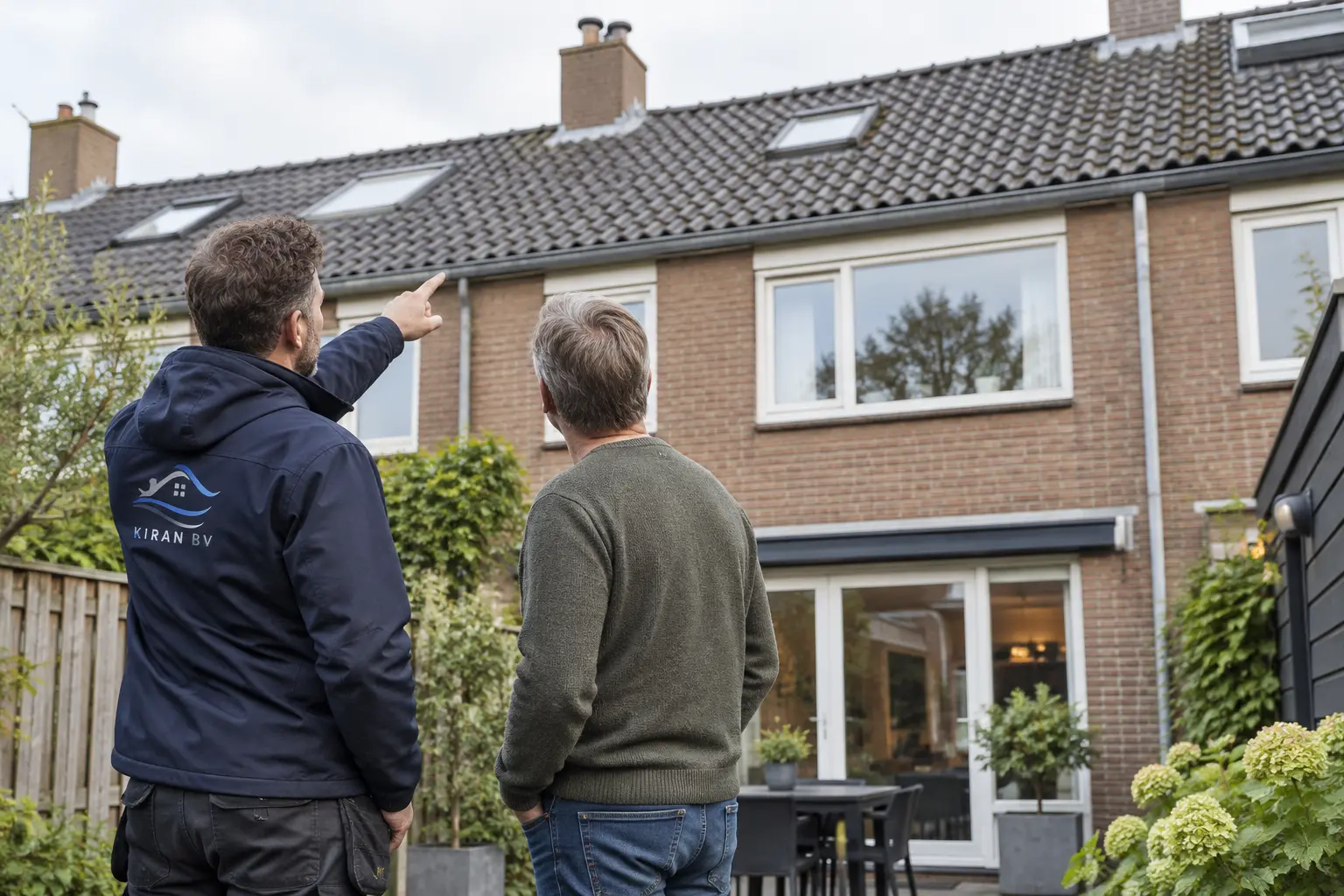 Een dakdekker en een bewoner kijken vanaf de tuin omhoog naar het dak van de woning